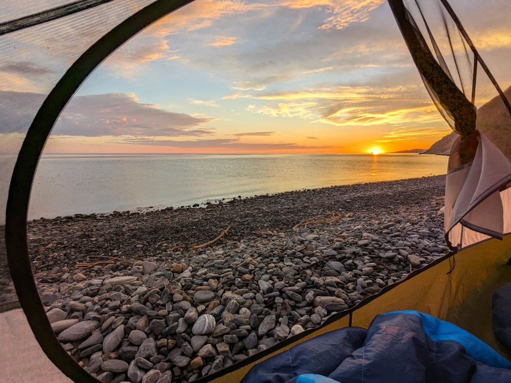 Looking through the door of a tent while camping on a beach. A bright sunrise is in the background.