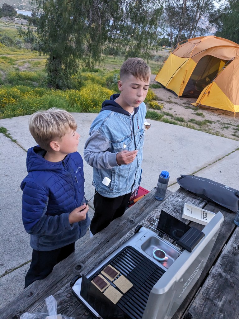 Two children cooking s'mores over a stove with a tent in the background.